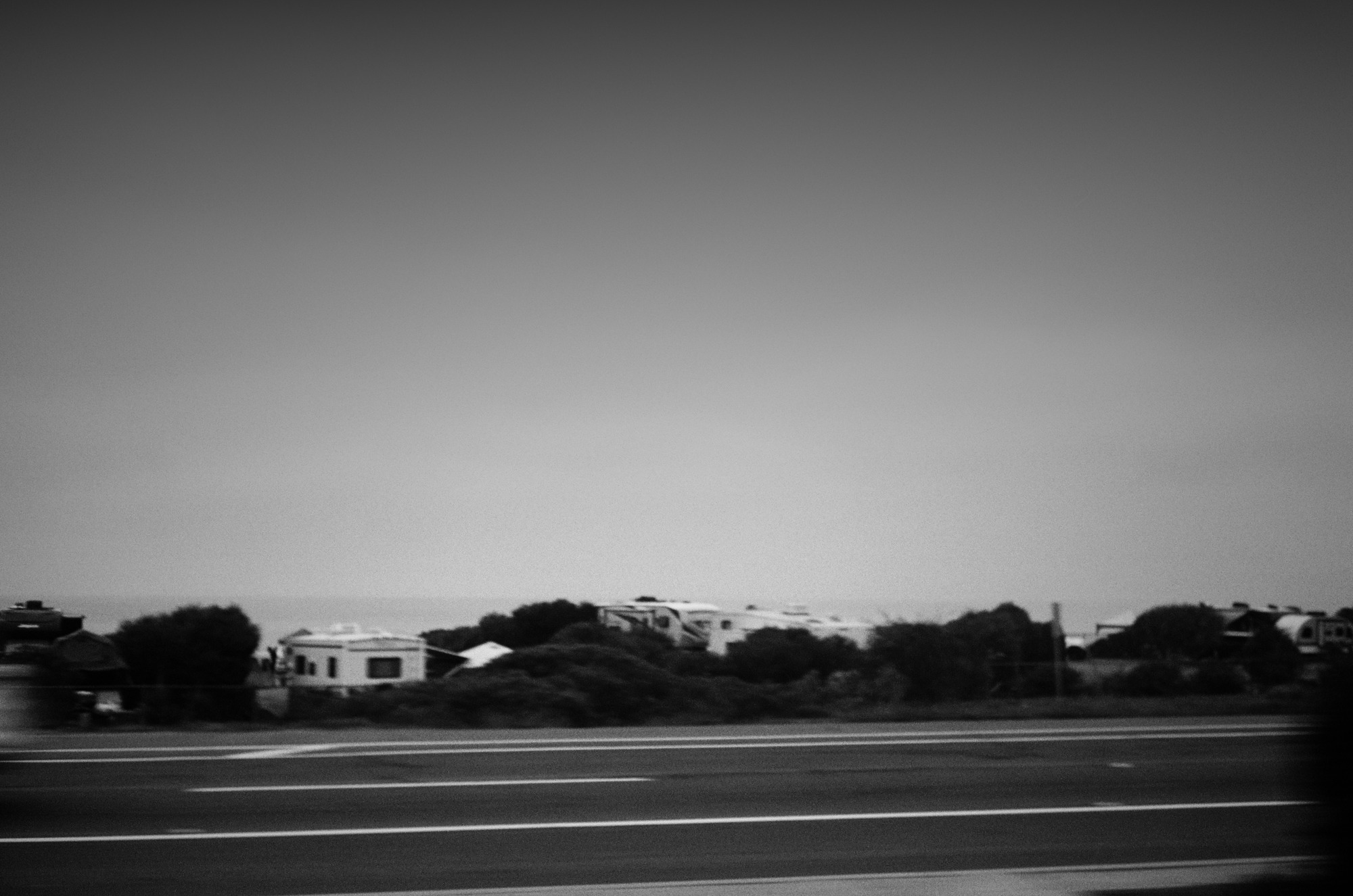 A coastal road in black and white, with bright lane markings and pale campers below a dark graduated sky.